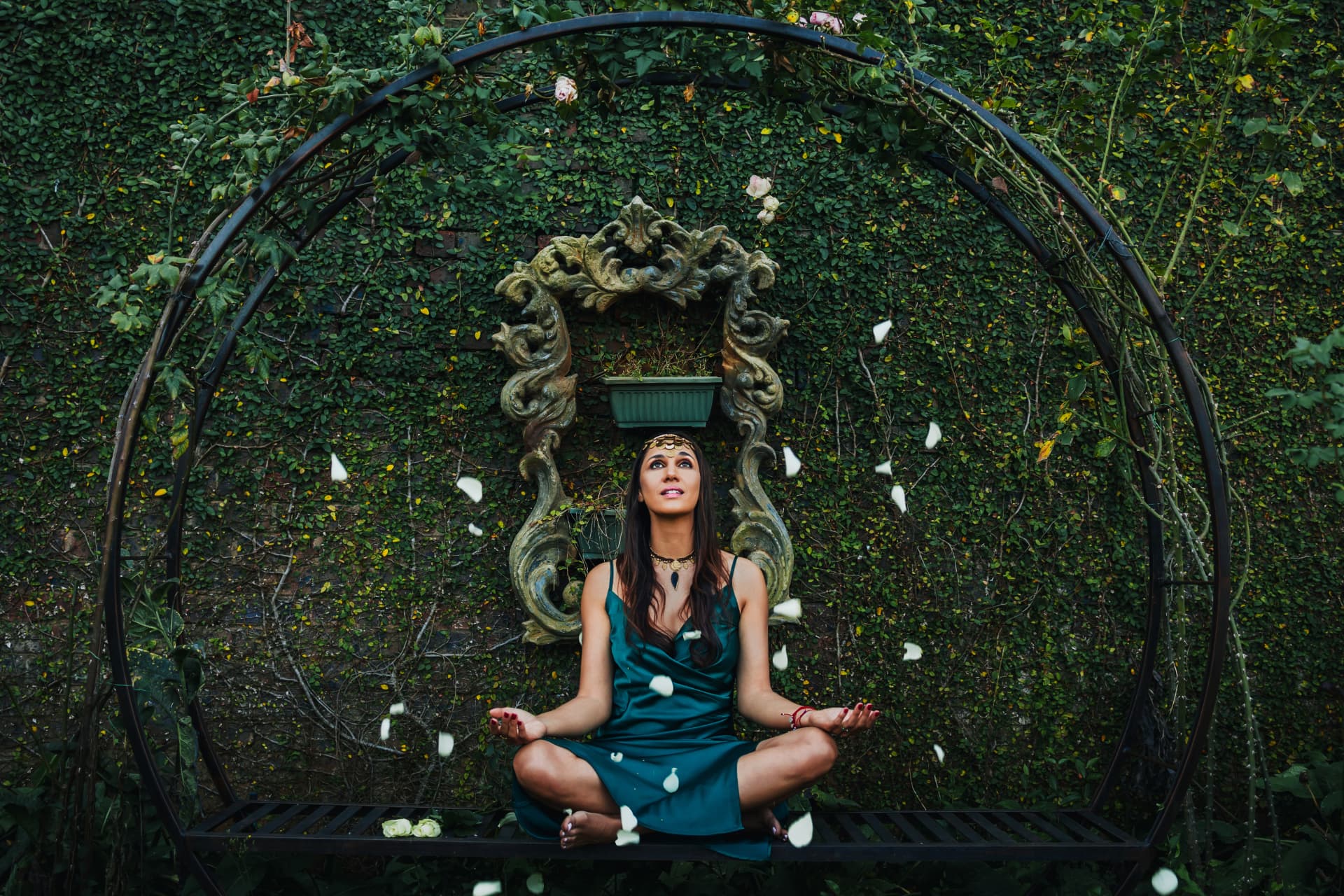 Noa seated in a garden arch with white rose petals falling around her