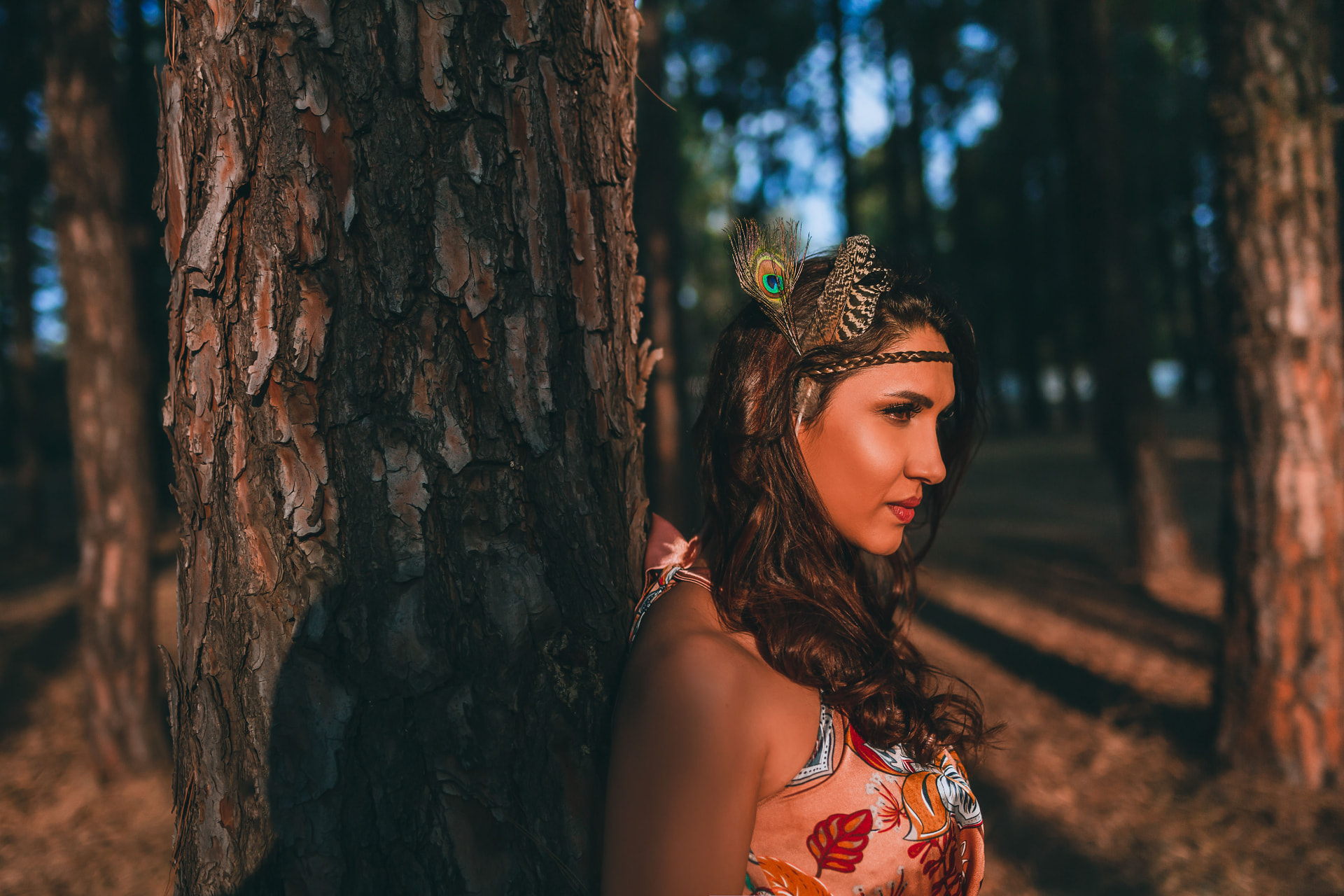 Noa resting against a pine trunk in golden light, feathers in her hair